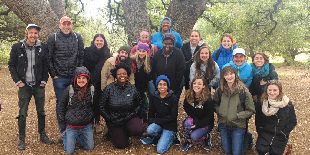 A group of people all dressed in winter clothing smile at the camera surrounded by live oak trees.