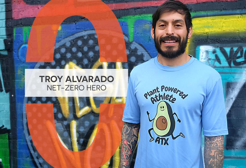 Troy Alvarado smiles at the camera while standing in front of a colorful mural. He is wearing a blue shirt with an avocado cartoon that says, "Plant Powered Athlete ATX."