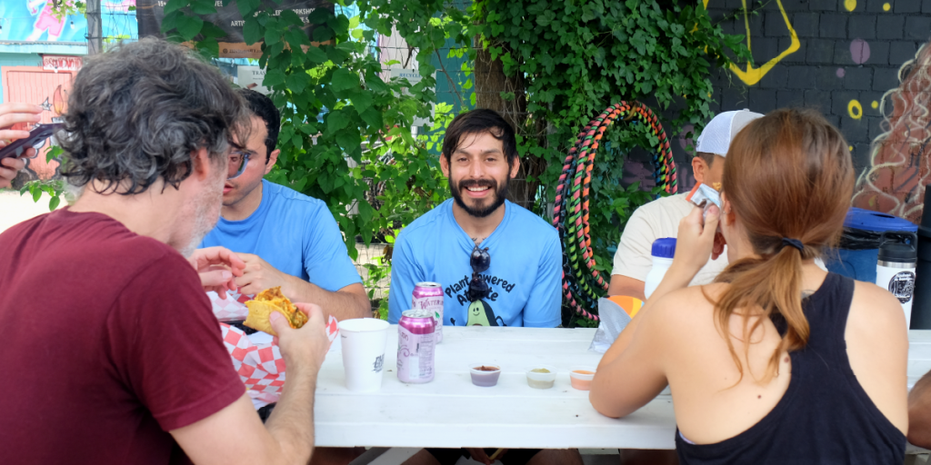 Troy smiles from the center of a picnic table as people eat vegan tacos around him.