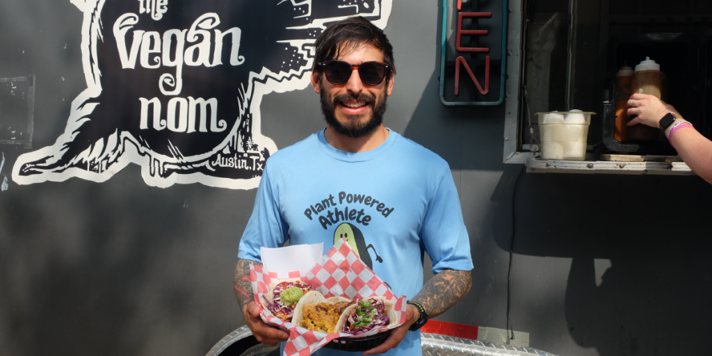Troy holds up a tray of tacos in front of The Vegan Nom food truck.