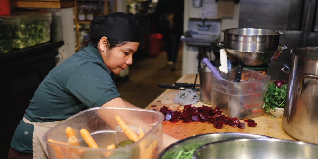 A woman pulls cut beets off of a large wooden countertop. She is surrounded by bins of vegetables.