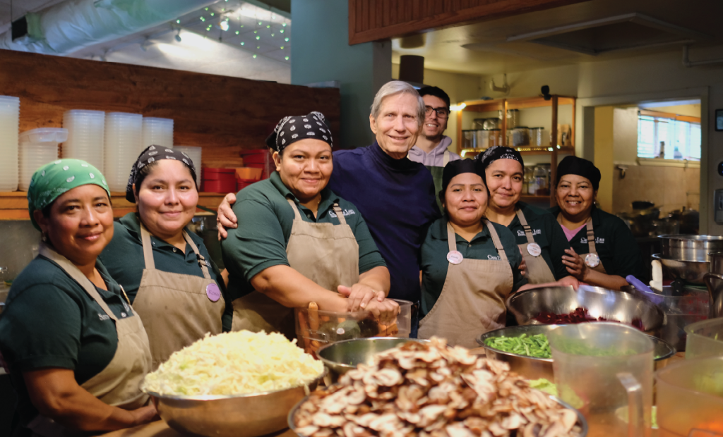 Wayo is surrounded by people in aprons in the kitchen of at Casa de Luz. Cut vegetables sit in large bowls on the counter in front of them.