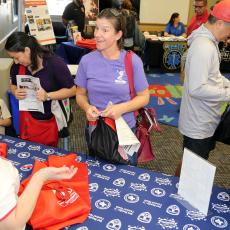 Caucasian woman smiling and speaking to another woman at a community outreach event with other people meeting and speaking to each other at tables.