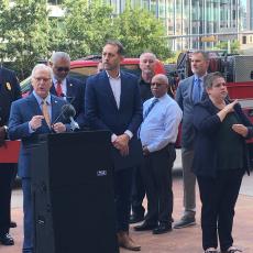 City of Austin Mayor Kirk Watson speaks at the podium during a press conference about wildfire danger. Other elected officials and fire chiefs stand behind him with 2 fire trucks in the background. A television camera is in the foreground.