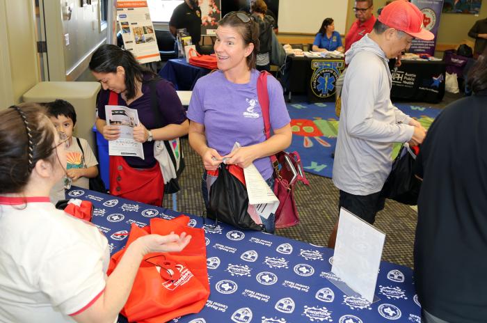 Caucasian woman smiling and speaking to another woman at a community outreach event with other people meeting and speaking to each other at tables.