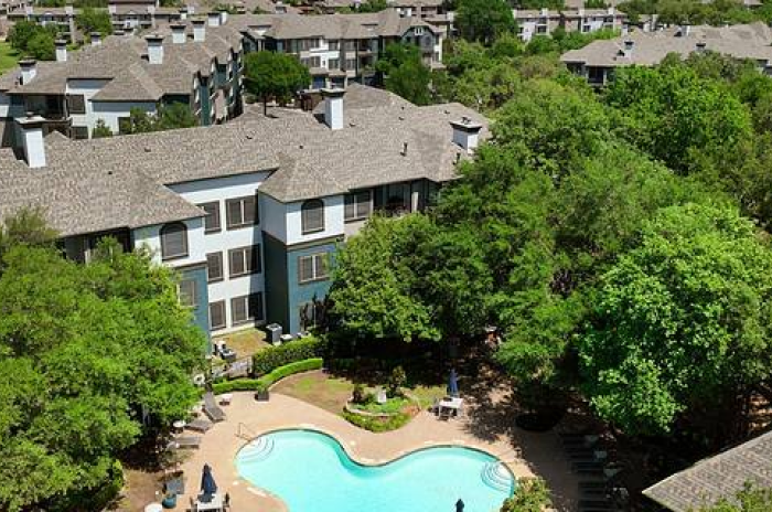 An aerial view of Twelve100, a residential complex, surrounded by lush green trees. The buildings have gray roofs and light-colored walls. A central courtyard features a large swimming pool with patio seating, creating a serene and inviting atmosphere.