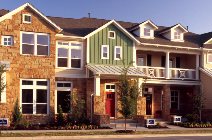 In Austin's Mueller neighborhood: A row of modern two-story townhomes with a mix of tan stone and light siding. The homes have large windows, red doors, and small landscaped gardens. Some parts of the buildings have green siding, and metal roofs cover the entryways. Young trees line the sidewalk, and signs in front of the homes display their names.