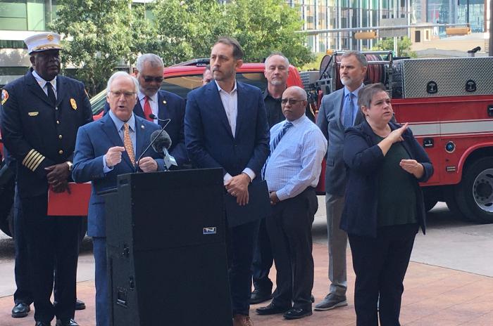 City of Austin Mayor Kirk Watson speaks at the podium during a press conference about wildfire danger. Other elected officials and fire chiefs stand behind him with 2 fire trucks in the background. A television camera is in the foreground.